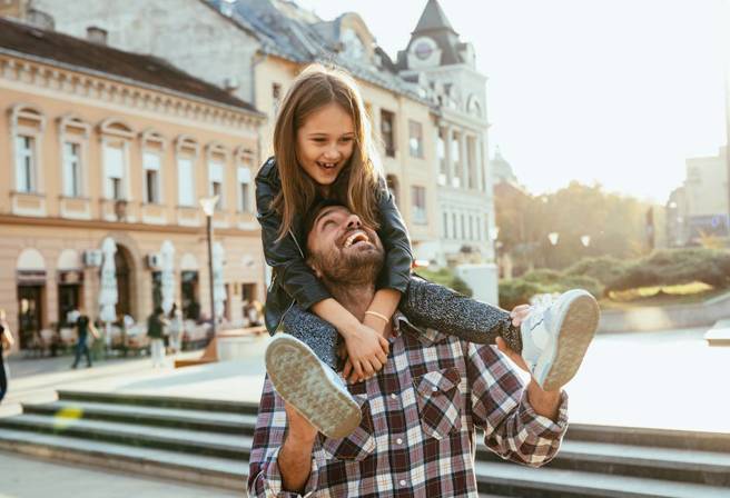 A father carries his daughter on his shoulders through a town square in a European city.